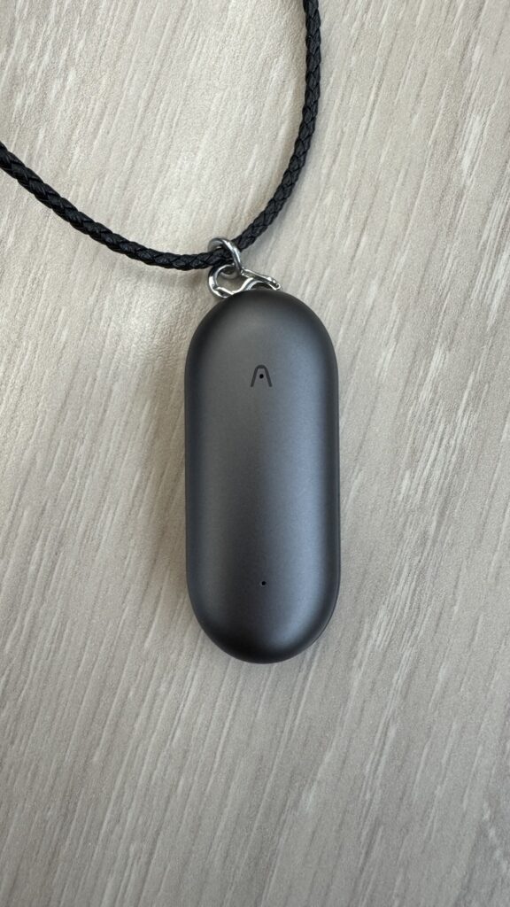 The Plaud Note Pin on a wooden table attached to the necklace attachment in dark grey/black colour